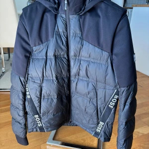 Svart pufferjacka från BOSS med huva - Svart pufferjacka från BOSS med avtagbara ärmar så den kan bäras som väst. Jackan har huva, dragkedja framtill och tydliga BOSS-loggor på banden längs sidorna. Materialet är en mix av polyester och syntet, perfekt för kyliga dagar.