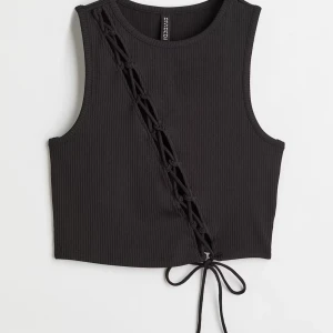 Svart ribbad linnetopp med snörning - Säljer en svart croppad linnetopp från H&M med ribbad struktur och snörningsdetalj i diagonal över framsidan. Toppen är ärmlös och har en snygg knytning längst ner.  Köpt för 149 men inte använd.