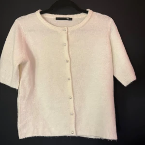 Vit kofta med pärlknappar från 157 - Superfin vit kofta från 157 med korta ärmar och rund hals. Koftan är stickad i mjukt material och har snygga pärlknappar framtill. Perfekt att slänga över en topp för en söt och stilren look.