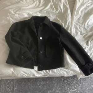 Svart kappa - Stilren svart jacka i ull med klassisk krage och tre stora knappar framtill. Jackan har två fickor framtill och långa ärmar med knappdetalj vid ärmslut. Perfekt för kyligare dagar och enkel att matcha med olika outfits.