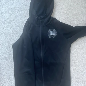 Svart hammarby hoodie  - Svart hoodie från hammarby med hel dragkedja och huva. Tryck med emblem på vänster bröst. Snygg och enkel design, perfekt för chill dagar eller när du ska på match. Långärmad och i mjukt material.