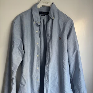 Blå skjorta från Ralph Lauren - Klassisk ljusblå skjorta från Ralph Lauren  med broderad logga på bröstet. Skjortan har knappar hela vägen och button down-krage. Perfekt till jeans eller chinos för en clean och stilren look. Inga defekter skick 9/10