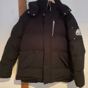 Svart dunjacka från Moose Knuckles - Säljer en svart dunjacka från Moose Knuckles med ikonisk logga på ärmen och snygga silvriga dragkedjor. Jackan har hög krage, stora fickor framtill och medföljande pälskrage. Perfekt för kalla dagar och riktigt stilren look.