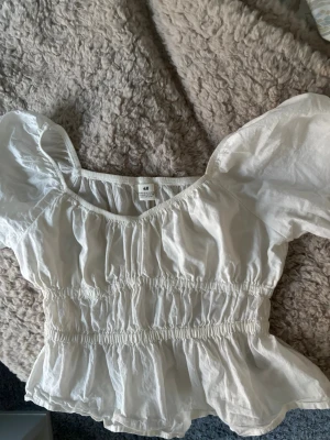 Vit blus med puffärmar från H&M - Säljer en vit blus från H&M med puffiga korta ärmar och smockad midja. Blusen har en rundad halsringning och volang nedtill, vilket ger en romantisk och trendig look. Perfekt för dig som gillar feminina detaljer och ljusa färger.