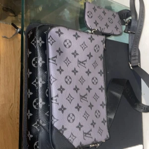  axelväska monogram svart/grå - Säljer en snygg axelväska i svart och grått med klassiskt monogrammönster. Väskan har justerbar axelrem, flera fack och en mindre avtagbar väska. Tillverkad i skinn med silverfärgade detaljer och dragkedja. Perfekt för dig som vill ha en stilren och lyxig look.