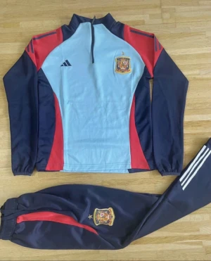 Adidas Spanien träningsställ blå/röd - Säljer ett Adidas träningsställ för Spanien med jacka och byxor. Färgerna är ljusblå, marinblå och röd med vita Adidas-ränder på byxbenet. Jackan har halv dragkedja och landslagets emblem på bröstet och byxan. Perfekt för fotbollsträning eller chill.