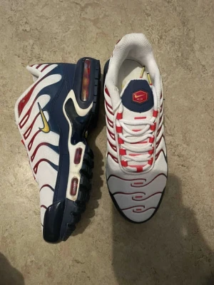 Nike Air Max Plus Tn sneakers vit/blå/röd - Nike Air Max Plus Tn sneakers i vitt med röda och blå detaljer, gul swoosh och Tn Air-logga på hälen. Skorna har snörning, mesh-överdel och synlig Air Max-dämpning i sulan. Snyggt randigt mönster och platt sula för en sportig vibe.