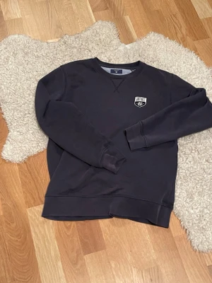 Mörkblå sweatshirt från GANT - Säljer en mörkblå sweatshirt från GANT med broderad logga på bröstet. Tröjan har rund hals, ribbade muddar vid ärmslut och nederkant samt långärmad passform. Perfekt för dig som gillar klassisk och enkel stil.                                                                                                                   OBS:det syns inte på bilden men den har ”armbågsskydd”. Be om bild på det. 