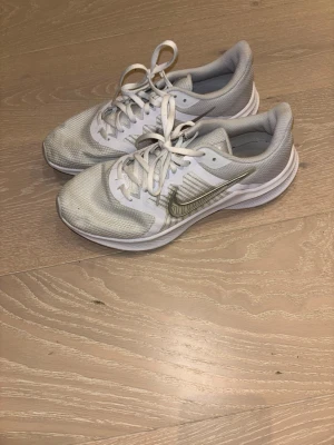 Nike Downshifter vita sneakers - Nike Downshifter sneakers i vitt och ljusgrått med mesh-ovandel och klassisk Nike-logga i silver på sidan. Skorna har snörning och platt sula, perfekt för dig som gillar sportig stil. Lätta och ventilerande med syntetmaterial och textilfoder.