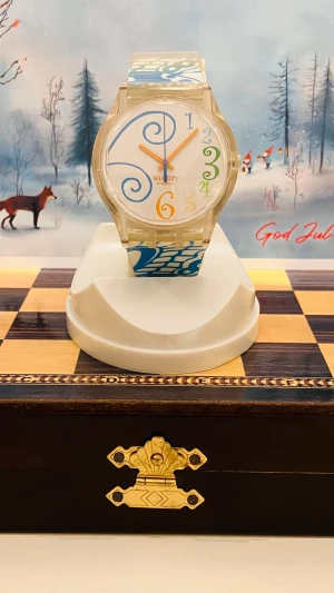 Swatch klocka med färgglada siffror - Unik Swatch armbandsklocka med transparent boett och vitt armband dekorerat med blå, gröna och gula grafiska mönster. Urtavlan har stora, snirkliga siffror i blått, grönt och gult samt orange visare. Klockan är i plast och har en lekfull, modern design.