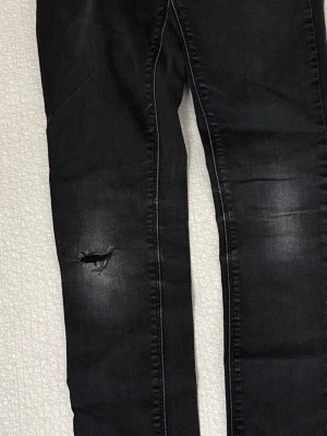 Svarta high waist Harper jeans Cubus - Svarta stretchiga jeans från Cubus, modell High Waist Harper. Skinny fit med hög midja och klassisk femficksdesign. Jeansen har en snygg sliten detalj vid knät och är tillverkade i bomull med stretch för extra komfort.