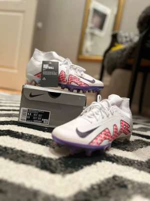 Nike Mercurial Zoom Superfly 9 Elite AG - Nike Mercurial Zoom Superfly 9 Elite AG fotbollsskor i vitt med lila sula och röda grafiska detaljer. Skorna har snörning, hög strumpkrage och en platt sula med dobbar för konstgräs. Materialet är syntet och designen är riktigt sportig med coola mönster längs sidorna.