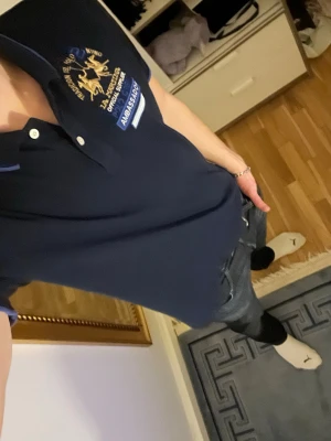 Mörkblå pikétröja La Martina M - Säljer en mörkblå pikétröja från La Martina i storlek M. Tröjan har broderade detaljer med guld och blått på bröstet, samt texten 'AMBASSADOR'. Klassisk krage med två knappar och tunna blå kanter på krage och ärmslut. Perfekt för en sportig och stilren look.