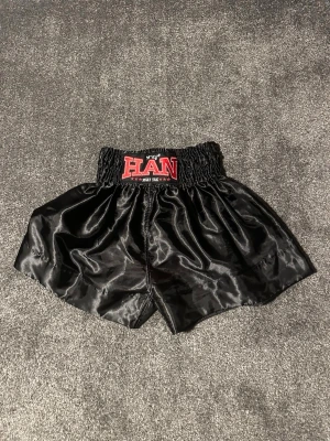 Svarta Muay Thai shorts från HAN - Säljer ett par svarta Muay Thai shorts från HAN i storlek S. De har hög midja med bred resår och är tillverkade i glansigt syntetmaterial. Klassisk boxningslook med röd och vit logga framtill. Perfekta för träning eller kampsport.