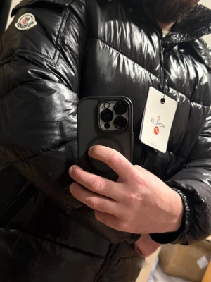 Svart pufferjacka från Moncler - Säljer en svart pufferjacka från Moncler med glansig finish och klassisk logga på ärmen. Jackan har huva, dragkedja framtill och praktiska fickor. Fylld med dun för extra värme och har snygga quiltade paneler. Perfekt för kalla dagar och riktigt stilren look.