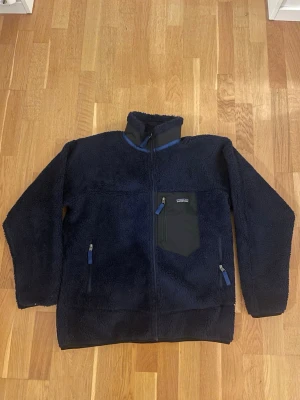 Mörkblå teddyjacka från Patagonia - Mysig mörkblå teddyjacka från Patagonia med hög krage och tre fickor med dragkedja. Jackan har en kontrasterande svart bröstficka och blå detaljer på dragkedjorna. Perfekt för kalla dagar och chill stunder utomhus. Aldrig använd