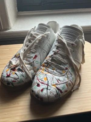Maison Margiela sneakers med färgstänk - Unika vita sneakers från Maison Margiela med färgstänk i rött, gult, svart och grönt. Skorna har snörning, rund tå och platt sula. Tillverkade i skinn med konstnärligt mönster som ger en cool vibe. Perfekt för dig som vill sticka ut med din stil.