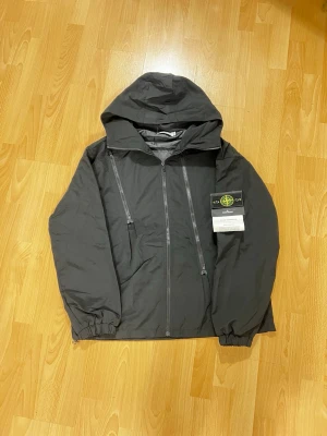 Svart vindjacka från Stone Island XL - Svart vindjacka från Stone Island med huva och två vertikala dragkedjor framtill. Jackan är tillverkad i polyester med Raso Gommato-ytbehandling som gör den vatten- och vindresistent. Klassisk Stone Island-logga på vänster ärm. Perfekt för dig som gillar streetwear.