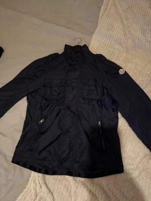 Moncler Field jacka - Säljer en mörkblå vindjacka från Moncler med hög krage, fyra framfickor med lock och knappar samt två sidofickor med dragkedja. Jackan har Moncler-logga på ärmen och snygga axeldetaljer. perfekt för vår och höst.