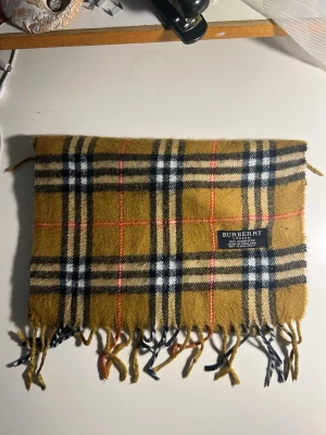 Rutig Burberry väst i kashmir - Säljer en klassisk rutig väst från Burberry i 100% kashmir. Färgen är beige med svarta, vita och röda rutor. Västen har fransar längs nederkanten och är tillverkad i England. Perfekt för dig som gillar tidlös design och mjuka material.