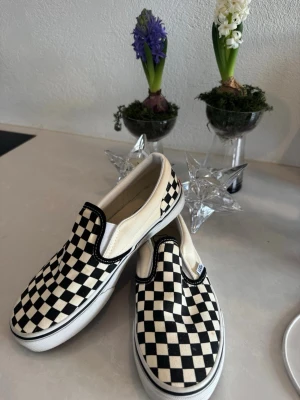 Vans slip-on checkerboard sneakers - Säljer ett par klassiska Vans slip-on sneakers med svart och vit rutig checkerboard-mönster. Skorna har en platt sula i gummi och är tillverkade i bomullstyg. Ikonisk röd Vans-logga bak på hälen och elastiska sidor för enkel påtagning.