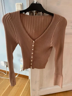 Beige ribbad kofta med pärlknappar - Snygg beige ribbad kofta med v-ringning och dekorativa pärlknappar framtill. Modellen är croppad med långärmade smala ärmar och har en mjuk, stickad känsla. Perfekt för dig som gillar stilrena detaljer och vill ha en kofta som sticker ut lite extra.
