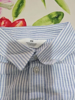 Blårandig skjorta från H&M - Snygg skjorta från H&M med blå och vita vertikala ränder. Klassisk krage, långärmad och med knappar framtill. Skjortan har en bröstficka och är gjord i mjuk bomull som känns skön mot huden. Perfekt för dig som gillar en clean och fräsch stil.