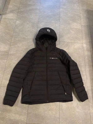 Svart pufferjacka från Sail Racing - Säljer en svart pufferjacka från Sail Racing med huva och dragkedja framtill. Jackan har två sidofickor med dragkedja och disk soplogga på bröstet och huvan. Perfekt för kalla dagar och riktigt skön passform. 
