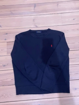 Mörkblå stickad tröja Polo Ralph Lauren - Snygg mörkblå stickad tröja från Polo Ralph Lauren med klassisk rund hals och röd broderad logga på bröstet. Tröjan har långa ärmar och ribbade muddar vid ärmslut och nederkant. Perfekt för dig som gillar stilrena och tidlösa plagg.