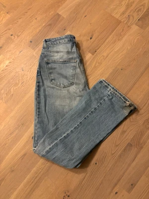 Gina tricot ljusblå bootcut jeans - Säljer ett par klassiska gina jeans i ljusblå tvätt med bootcut-ben. Jeansen har fyra fickor, slitning vid knät och råa kanter längst ner. Tillverkade i jeansmaterial av bomull, perfekt för dig som gillar retrostil och avslappnad vibe.
