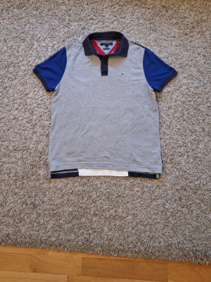Tommy Hilfiger flerfärgad pikétröja  - Snygg pikétröja från Tommy Hilfiger i storlek S(passar M också). Använd fåtal gånger och skicket är riktigt bra, inga slitningar eller defekter finns. Den har en grå framsida, blå ärmar och krage, samt en cool baksida med breda röda, vita och blå paneler. Skick: 9/10