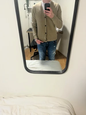 Beige kofta med knappar - Snygg beige kofta med svarta knappar och ribbade muddar från Massimo dutti. Perfekt att slänga på över en t-shirt för en chill och stilren look. Koftan har lång ärm och klassisk v-ringad design. Passar dig som gillar enkel och clean stil.