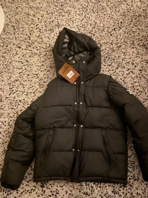 Svart  Burberry jacka med huva - Helt ny svart pufferjacka med huva Storlek: M Aldrig använd, taggar kvar. Jackan har avtagbara ärmar (kan även bäras som väst). Stilren design med rutigt foder i huvan. Passar bra för någon som är ca 160–170 cm och väger 50–70 kg. Varm och perfekt för vintern.