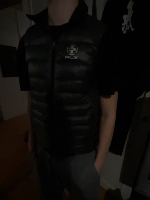 Svart dunväst från RLX - Säljer en svart dunväst från RLX med hög krage och dragkedja framtill. Västen har ett stilrent, quiltat mönster och RLX-logga på bröstet. Perfekt att slänga över en hoodie eller t-shirt när det är kyligt ute. Fickor med dragkedja på sidorna.