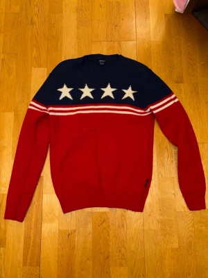 Röd och marinblå stickad tröja GANT - Snygg stickad tröja från GANT i rött och marinblått med vita stjärnor och ränder över bröstet. Tröjan har rund hals och långa ärmar. Materialet är 80% lammull och 20% polyamid, vilket gör den både mjuk och varm. Perfekt för dig som vill sticka ut med cool design.