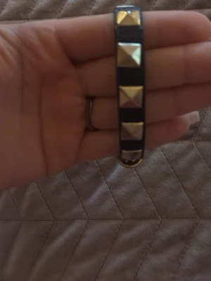 Svart skinnarmband med nitar - Snyggt svart armband i skinn med guldiga pyramidformade nitar och spänne. Perfekt accessoar för att ge din outfit lite edge. Justerbar längd med klassiskt bältesspänne i metall.