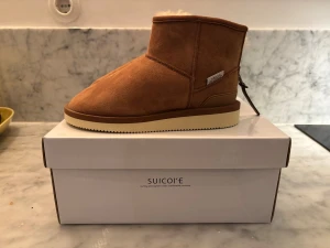 Suicoke ELS-M2ab lo storlek 35 - Säljer ett par bruna boots från Suicoke med mjukt foder och rund tå. Skorna är tillverkade i mocka med beige platt sula och har en låg, klassisk siluett. Perfekta för kalla dagar med sin fluffiga insida som håller fötterna varma.