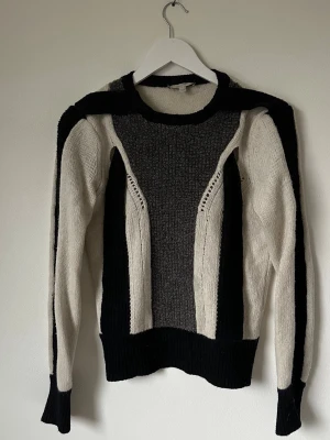 Svart och grå stickad tröja Dagmar XS - Unik stickad tröja från Dagmar i storlek XS med coolt grafiskt mönster i svart, grått och beige. Materialet är 65% lammull och 35% nylon, vilket gör den både mjuk och varm. Tröjan har långärmad passform och ribbade muddar vid ärmslut och nederkant. 5L4