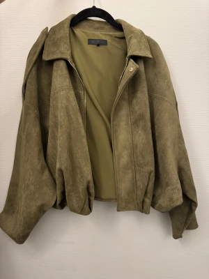 Olivgrön mockajacka från ZARA - Säljer en oversized olivgrön mockajacka från ZARA med bred krage och dragkedja framtill. Jackan har långa, vida ärmar och en mjuk, matt yta som ger en cool vintagekänsla. Perfekt för dig som gillar avslappnad och trendig stil.