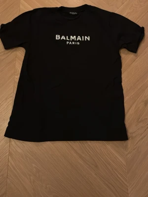 Svart Balmain Paris t-shirt - Svart t-shirt från Balmain Paris med vit logotyptryck på bröstet. Klassisk rund hals och korta ärmar. Materialet känns mjukt och skönt, perfekt för dig som gillar stilrena designer och vill ha något som sticker ut med en lyxig vibe.