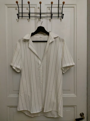 Vit randig skjorta med kort ärm - Snygg vit skjorta med tunna svarta ränder och klassisk krage. Skjortan har korta ärmar och knäppning framtill. Passar perfekt till jeans eller shorts för en chill look. Materialet känns lätt och luftigt, perfekt för varmare dagar.