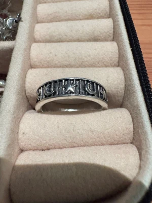 Silvrig ring med måne och stjärnor - Säljer en cool silvrig ring med ingraverade månar och stjärnor runt hela bandet. Ringen har en lite bredare form och en mörkare bakgrund som framhäver detaljerna. Perfekt för dig som gillar unika accessoarer med lite mystisk vibe.