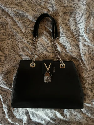 Svart handväska med kedjehandtag - Snygg svart handväska från Valentino med silverfärgade kedjehandtag och ett V-emblem framtill. Väskan är tillverkad i skinn och har en stilren, fyrkantig form. Perfekt för dig som gillar klassisk design med en modern twist. Väskan är aldrig använd!!🥰 Nypris var något runt 2000kr.