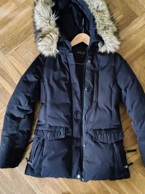 Svart parkas från Hollies med fuskpäls - Säljer en svart parkas från Hollies med stor huva kantad av fuskpäls. Jackan har dragkedja och knappar framtill, stora fickor och justerbar midja. Perfekt för kalla dagar och riktigt snygg till vintern.