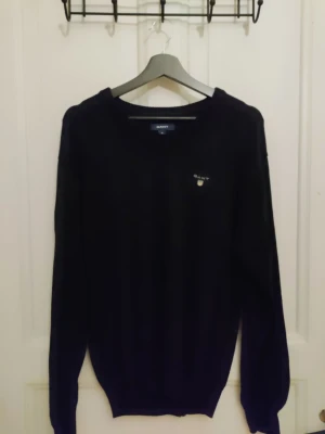 Svart stickad tröja från GANT - Svart stickad tröja från GANT med v-ringad hals och broderad logga på bröstet. Tröjan har långa ärmar och ribbade muddar vid hals, ärmslut och nederkant. Perfekt för dig som gillar klassisk och stilren design.