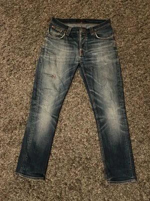 Nudie jeans blå tapered - Säljer ett par vintage Nudie jeans i blå tvätt som inte går att köpa längre från hemsidan med snygga slitningar och broderad bakficka. Modellen är tapered med normal passform och har en röd lagning på ena benet. Klassisk femficksdesign och tillverkade i jeansmaterial med lite stretch.