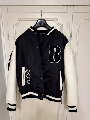 Svart och vit collegejacka från Zara - Säljer en svart och vit collegejacka från Zara i storlek M. Jackan har stora broderade detaljer, bland annat ett B på bröstet och en kedja med text på ryggen. Ärmarna är vita och kroppen svart, med ribbade muddar och tryckknappar framtill. 