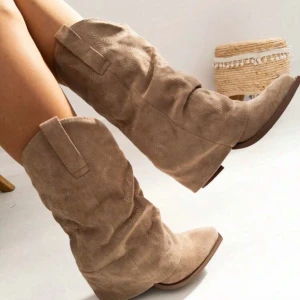 Beige cowboy boots i mocka - Säljer ett par snygga beige cowboy boots med spetsig tå och låg klack. Bootsens yta är i mjuk mocka med dekorativa sömmar och klassiska dragflikar på sidorna. Perfekt för dig som vill ha en cool westernstil.