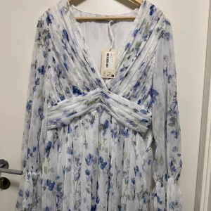 Blommig klänning från NA-KD - Säljer en superfin klänning från NA-KD med v-ringad omlottfront och blommigt mönster i blått, grönt och vitt. Klänningen har långa ärmar med volangdetaljer och ett vackert draperat midjeband. Perfekt för sommarens alla tillfällen. Passar stor M-XL