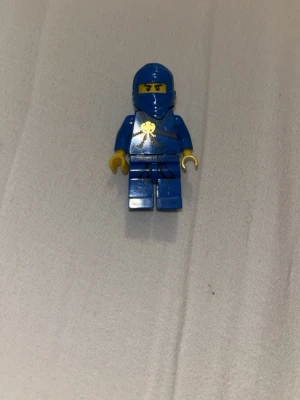 "OG Jay" - Lego Ninjago - Säljer en blå väst till LEGO-figur med coolt guldtryck framtill och svarta detaljer. Västen har en klassisk passform och är gjord i plast. Dock är han delvis skadad på ansiktet, men endå en perfekt för att ge din minifigur en ninja-look.